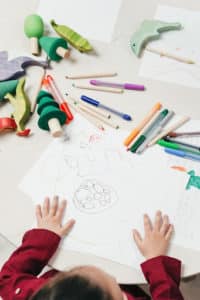 A child’s scribble drawing surrounded by pens and pencils on a table