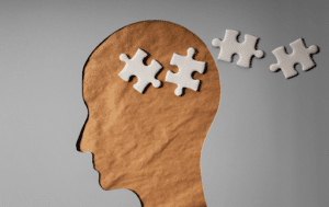 Papel gris recortado en forma de cabeza humana con piezas de puzzle en blanco donde estaría el cerebro.